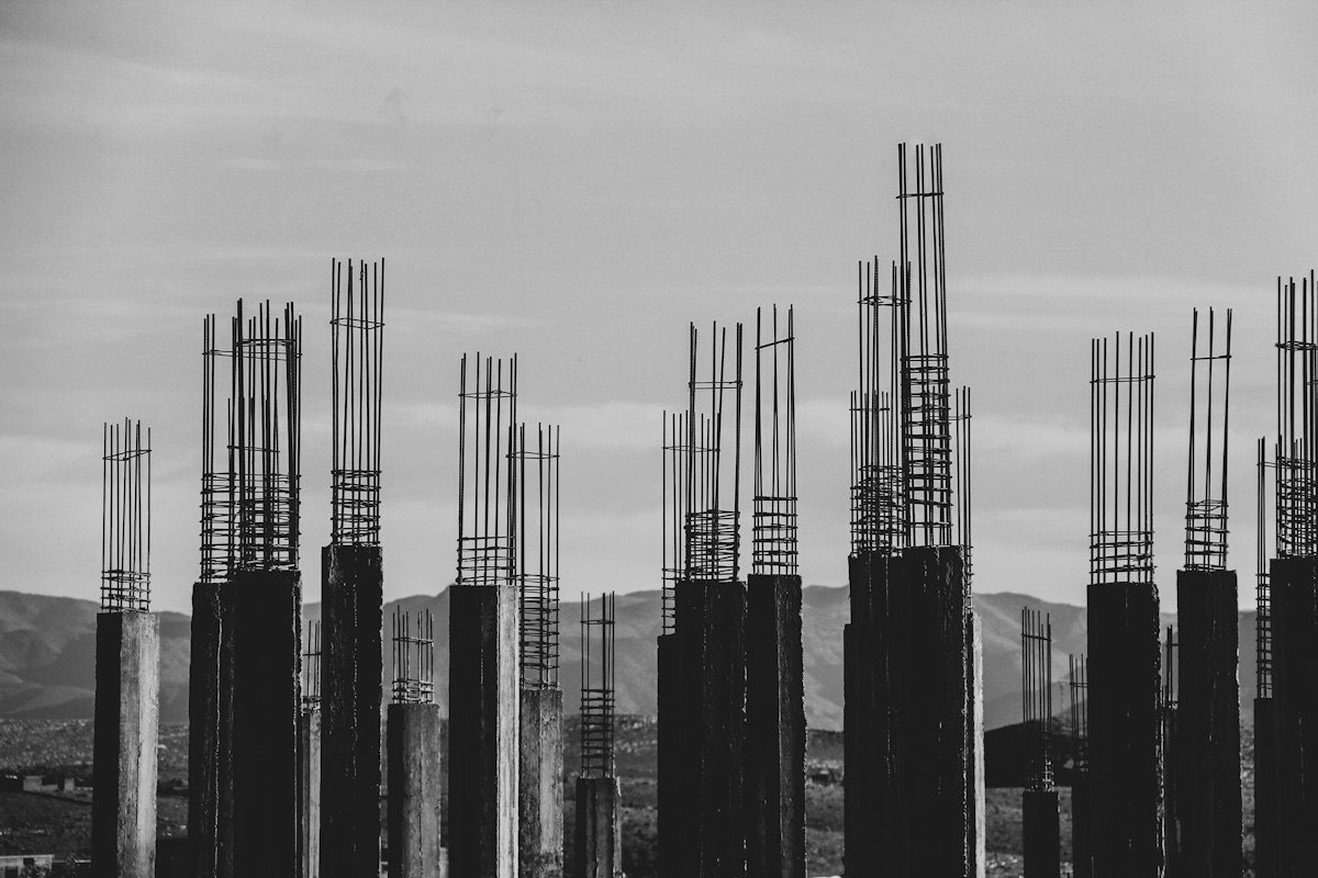 Schwarz-Weiß-Aufnahme von Betonstützen mit herausragender Bewehrung vor einer Hügellandschaft