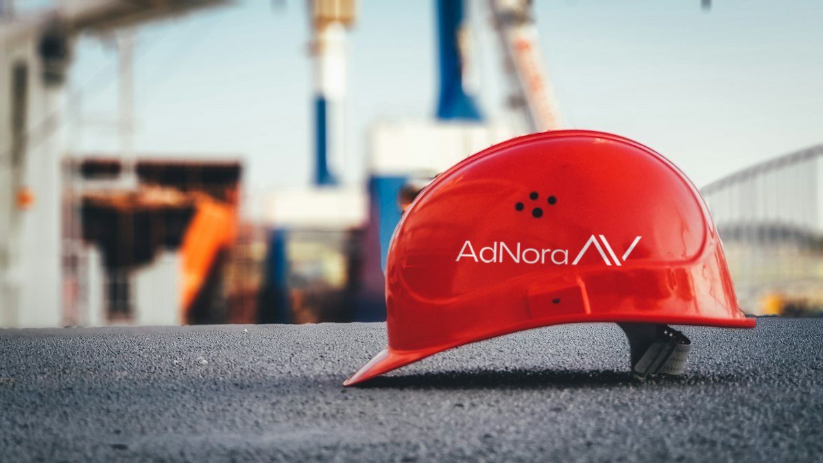 Roter Schutzhelm mit AdNora-Logo auf Asphalt vor unscharf erkennbaren Baukränen